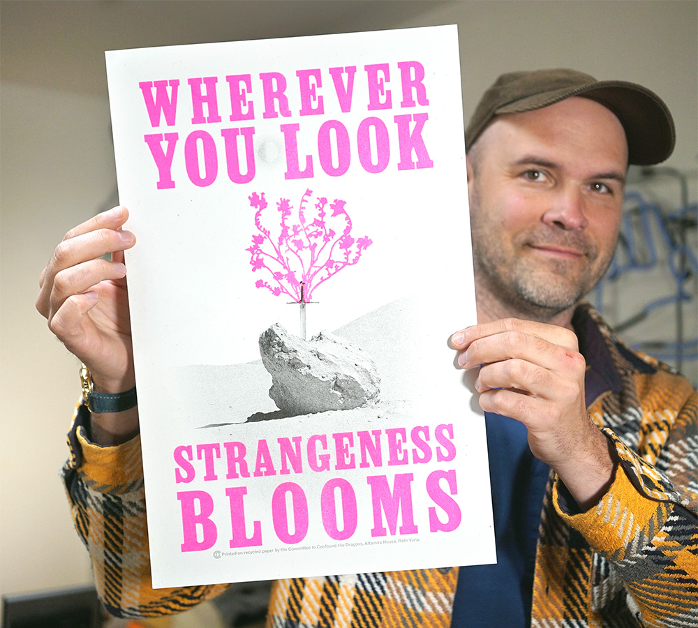 A picture of Robin holding up a tabloid-size zine, the letters printed in bold, bright pink ink.